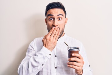 Young handsome man with beard drinking mate infusion beverage over white background covering mouth with hand, shocked and afraid for mistake. Surprised expression