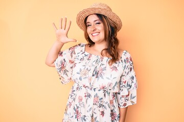 Young beautiful caucasian woman wearing summer dress and hat showing and pointing up with fingers number five while smiling confident and happy.