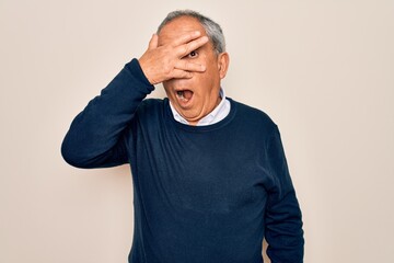 Senior handsome grey-haired man wearing sweater and glasses over isolated white background peeking in shock covering face and eyes with hand, looking through fingers with embarrassed expression.