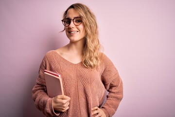 Young beautiful blonde woman wearing glasses holding notebook over pink background with a happy face standing and smiling with a confident smile showing teeth