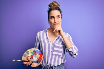 Young beautiful blonde artist woman painting using brush and palette over purple background serious...