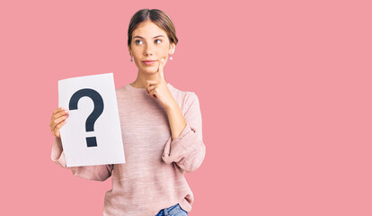 Beautiful caucasian woman with blonde hair holding question mark serious face thinking about question with hand on chin, thoughtful about confusing idea