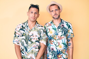 Young gay couple of two men wearing summer hat and hawaiian shirt relaxed with serious expression on face. simple and natural looking at the camera.