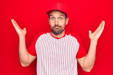 Naklejka premium Young handsome player man wearing baseball sportswear over isolated red background clueless and confused with open arms, no idea and doubtful face.