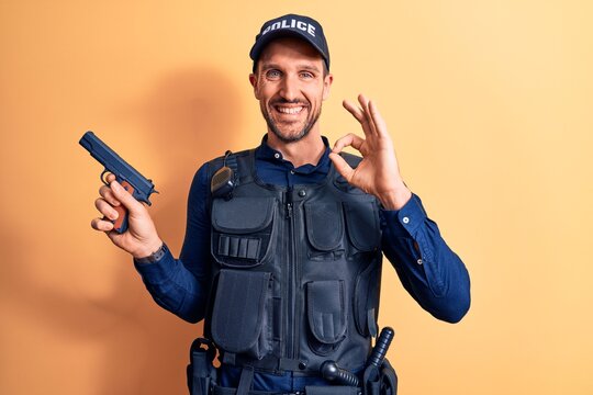Handsome policeman wearing uniform and bulletprof holding gun over yellow background doing ok sign with fingers, smiling friendly gesturing excellent symbol