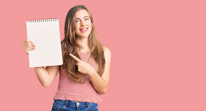 Beautiful caucasian young woman holding notebook smiling happy pointing with hand and finger