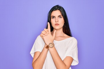 Young beautiful brunette woman wearing casual white t-shirt over purple background Holding symbolic gun with hand gesture, playing killing shooting weapons, angry face