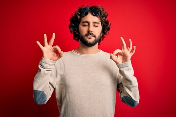 Naklejka premium Young handsome man with beard wearing casual sweater standing over red background relax and smiling with eyes closed doing meditation gesture with fingers. Yoga concept.