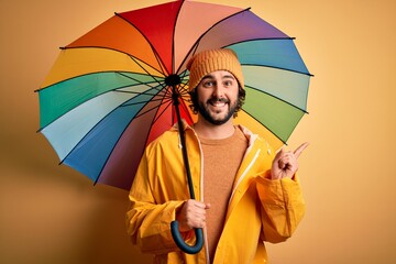 Young handsome man with beard wearing raincoat for rainy day holding colorful umbrella very happy pointing with hand and finger to the side