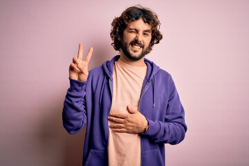 Young handsome sporty man with beard wearing casual sweatshirt over pink background smiling with happy face winking at the camera doing victory sign with fingers. Number two.