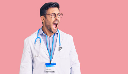 Young hispanic man wearing doctor uniform and stethoscope angry and mad screaming frustrated and...