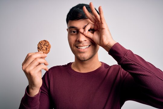 Young hispanic man eating chocolate chips cookie over isolated background with happy face smiling doing ok sign with hand on eye looking through fingers