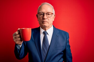 Senior grey haired business man drinking a hot cup of coffee over red background with a confident expression on smart face thinking serious