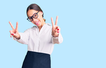 Beautiful brunette young woman wearing professional waitress apron smiling with tongue out showing fingers of both hands doing victory sign. number two.