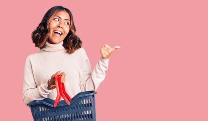 Young beautiful mixed race woman holding supermarket shopping basket pointing thumb up to the side smiling happy with open mouth