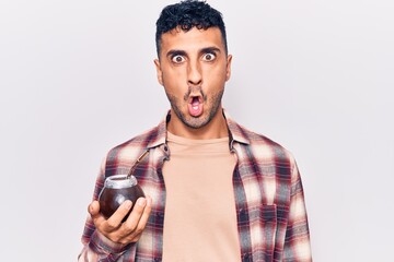 Young hispanic man drinking mate infusion scared and amazed with open mouth for surprise, disbelief face