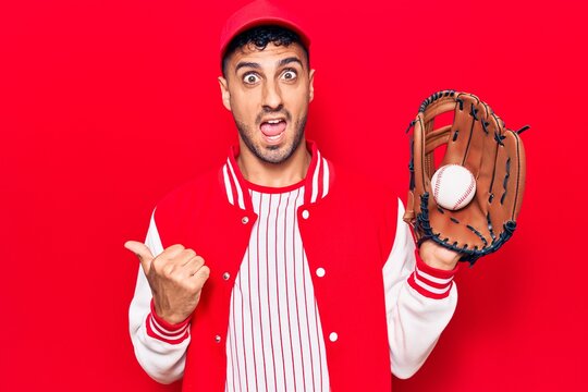 Young hispanic man wearing baseball uniform holding golve and ball pointing thumb up to the side smiling happy with open mouth