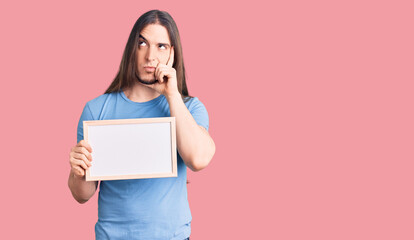 Young adult man with long hair holding empty white chalkboard serious face thinking about question...