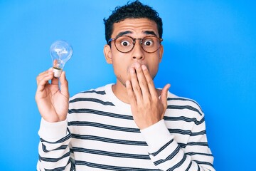 Young handsome hispanic man holding lightbulb for inspiration and idea covering mouth with hand, shocked and afraid for mistake. surprised expression