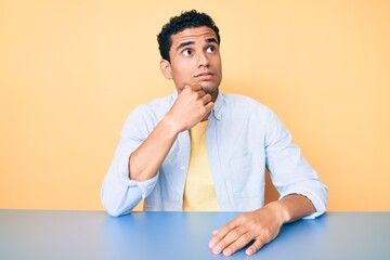 Young handsome hispanic man wearing casual clothes sitting on the table serious face thinking about question with hand on chin, thoughtful about confusing idea