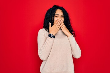 Young african american curly woman wearing casual turtleneck sweater over red background laughing and embarrassed giggle covering mouth with hands, gossip and scandal concept