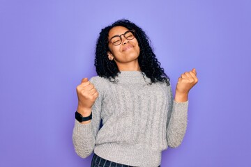 Young african american woman wearing casual sweater and glasses over purple background very happy and excited doing winner gesture with arms raised, smiling and screaming for success. Celebration
