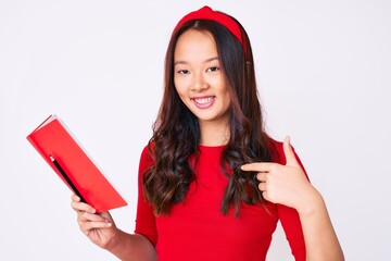 Young beautiful chinese girl holding book pointing finger to one self smiling happy and proud