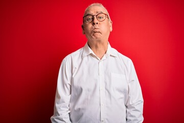 Middle age handsome hoary man wearing casual shirt and glasses over red background puffing cheeks with funny face. Mouth inflated with air, crazy expression.