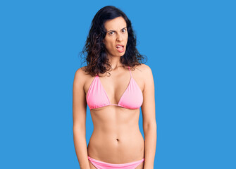Young beautiful hispanic woman wearing bikini in shock face, looking skeptical and sarcastic,...