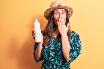 Young beautiful brunette woman wearing summer hat holding bottle of sunscreen lotion covering mouth...