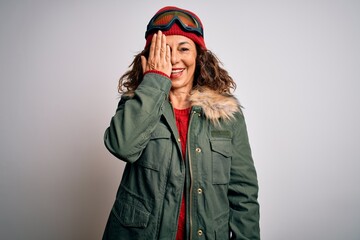 Middle age skier woman wearing snow sportswear and ski goggles over white background covering one eye with hand, confident smile on face and surprise emotion.