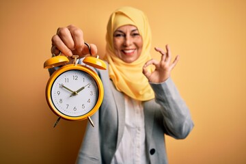 Middle age brunette business woman wearing muslim traditional hijab holding alarm clock doing ok sign with fingers, excellent symbol