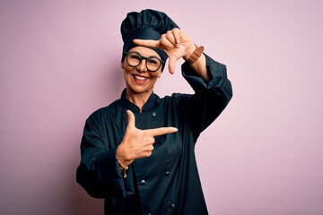 Middle age brunette chef woman wearing cooker uniform and hat over isolated pink background smiling making frame with hands and fingers with happy face. Creativity and photography concept.