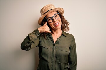 Middle age brunette woman wearing glasses and hat standing over isolated white background smiling doing phone gesture with hand and fingers like talking on the telephone. Communicating concepts.