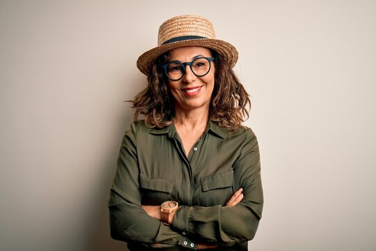 Middle age brunette woman wearing glasses and hat standing over isolated white background happy face smiling with crossed arms looking at the camera. Positive person.