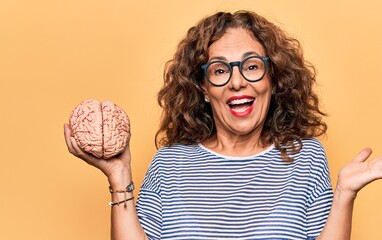Middle age brunette woman holding brain as mental care and memory health over pink background...