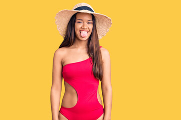 Young beautiful latin girl wearing swimwear and summer hat sticking tongue out happy with funny expression. emotion concept.