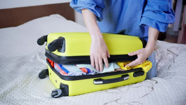 luggage for vacation, young woman is going to travel and trying to close her suitcase with things and clothes, sits on top and zips up on her travel bag while sitting on bed in room