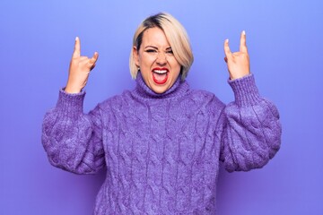 Beautiful blonde plus size woman wearing casual turtleneck sweater over purple background shouting with crazy expression doing rock symbol with hands up. Music star. Heavy music concept.