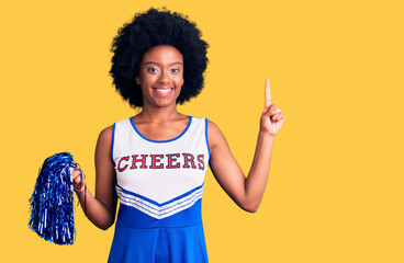 Young african american woman wearing cheerleader uniform holding pompom with a big smile on face, pointing with hand finger to the side looking at the camera.