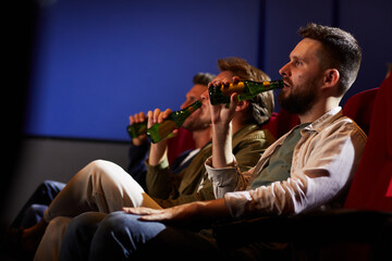 Side view at group of men watching movie in cinema and drinking beer while sitting in row on red...