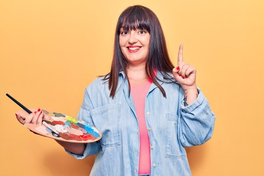Young plus size woman holding paintbrush and palette surprised with an idea or question pointing finger with happy face, number one