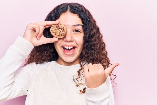 Beautiful kid girl with curly hair holding cookie pointing thumb up to the side smiling happy with open mouth
