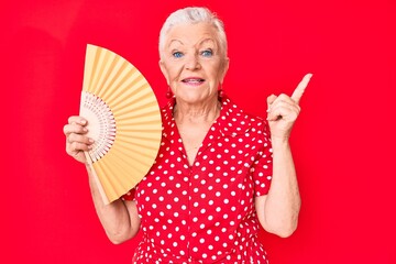 Senior beautiful woman with blue eyes and grey hair waving hand fan cooling air in summer smiling happy pointing with hand and finger to the side