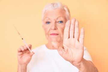 Senior beautiful woman with blue eyes and grey hair holding syringe with open hand doing stop sign with serious and confident expression, defense gesture