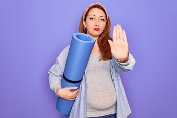 Young beautiful redhead pregnant woman expecting baby holding mat to do prenatal exercise with open hand doing stop sign with serious and confident expression, defense gesture
