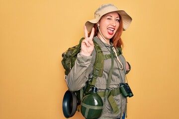 Young redhead backpacker woman hiking wearing backpack and hat over yellow background smiling with happy face winking at the camera doing victory sign with fingers. Number two.