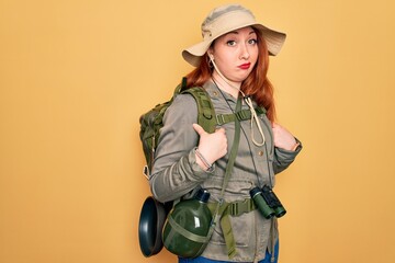 Young redhead backpacker woman hiking wearing backpack and hat over yellow background skeptic and...