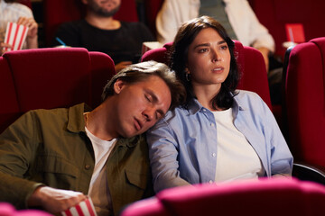 Portrait of young couple in cinema, focus on woman watching movie with bored boyfriend sleeping on...