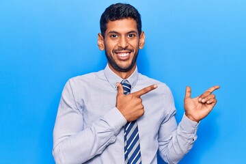 Young latin man wearing business clothes smiling and looking at the camera pointing with two hands and fingers to the side.